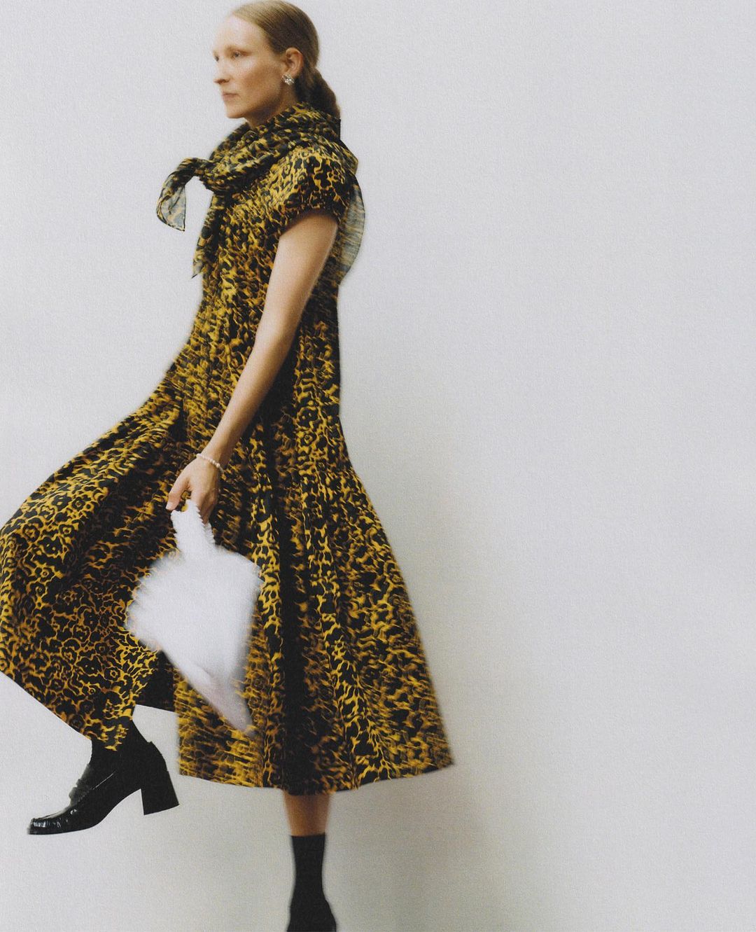 Fashionable woman in a leopard-print dress walks on a neutral background holding a white handbag. Her elegant style is emphasized.