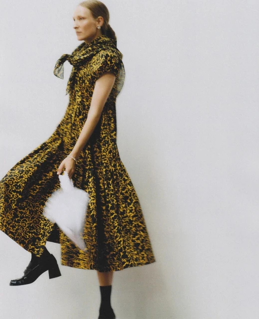 Fashionable woman in a leopard-print dress walks on a neutral background holding a white handbag. Her elegant style is emphasized.