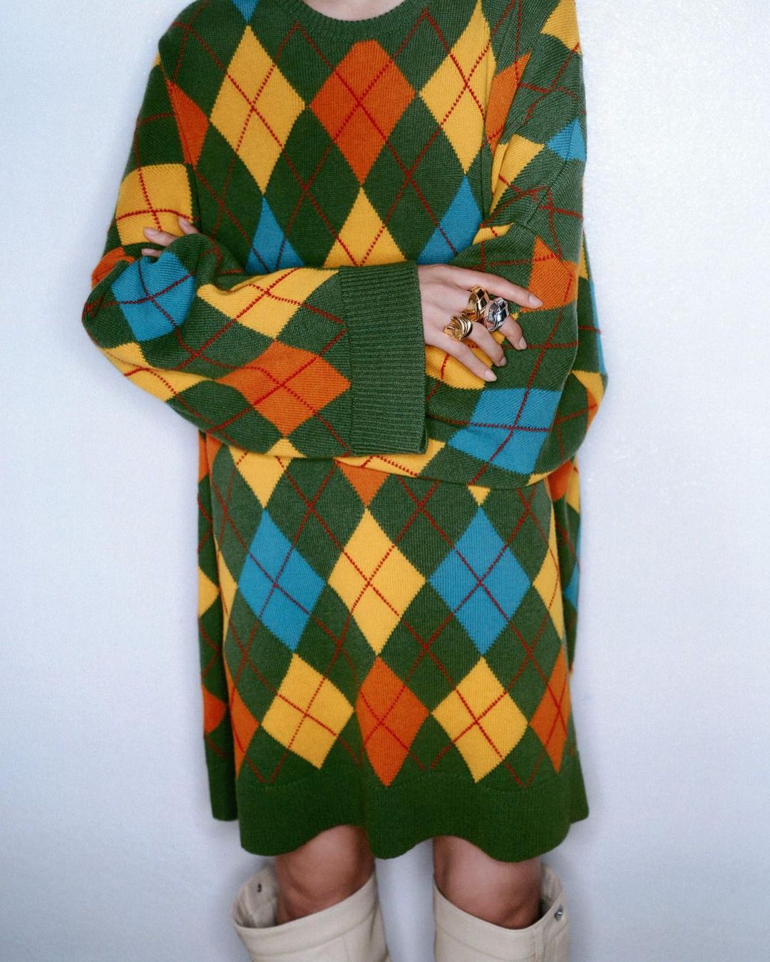 Woman wearing colorful argyle sweater and matching skirt, accessorized with large rings and cream-colored boots against white wall.