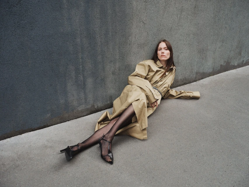 Adult woman in a tan trench coat poses while sitting on a grey concrete surface, against a mottled blue-grey wall, wearing black stockings and heels.