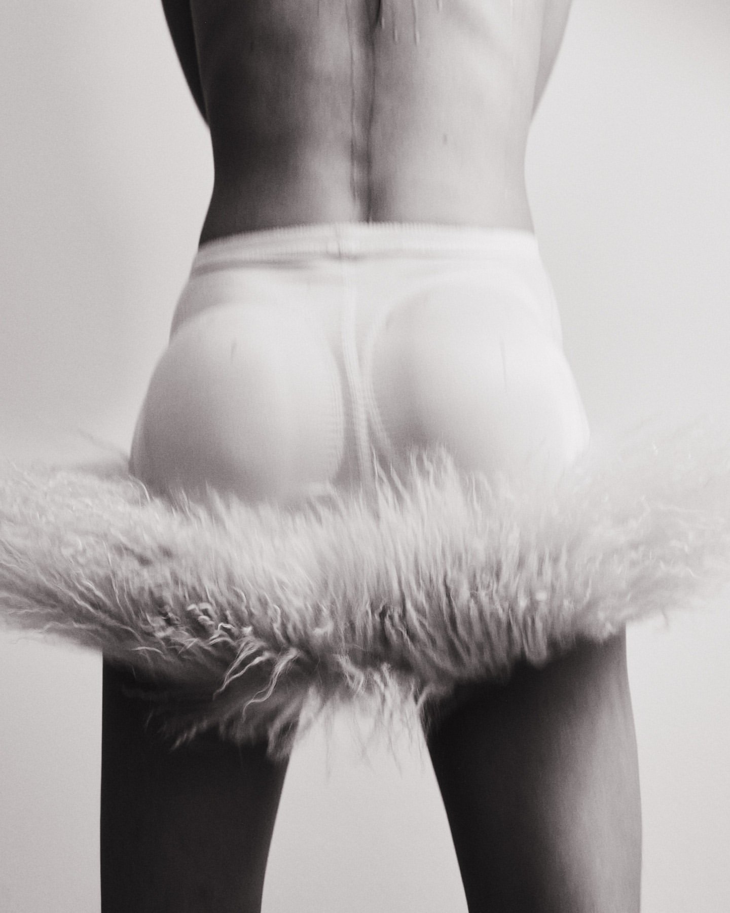 Back view of a person in a skirt with fur trim, captured in a black and white photo with a seamless white backdrop in a photo studio.