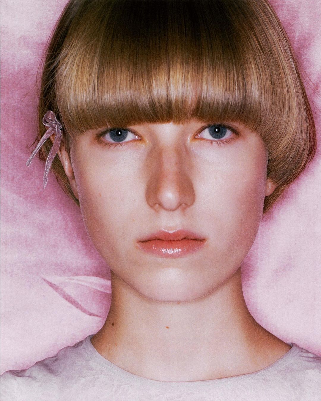 Close-up portrait of a young woman with a symmetrical haircut, blue eyes, and pale skin against a light pink background, wearing a light top.