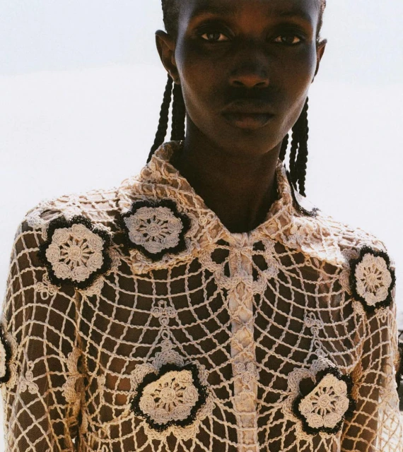 Close-up of an African-descent adult female model wearing a cream-colored, flower-decorated, crocheted blouse against a light outdoor background.