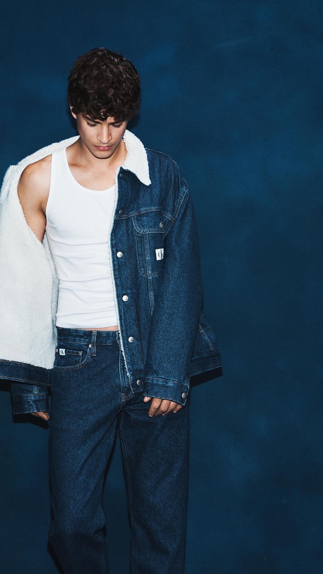 A young adult male poses in a white tank top and denim sherpa jacket and jeans in front of a dark blue studio backdrop.