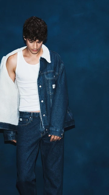 A young adult male poses in a white tank top and denim sherpa jacket and jeans in front of a dark blue studio backdrop.
