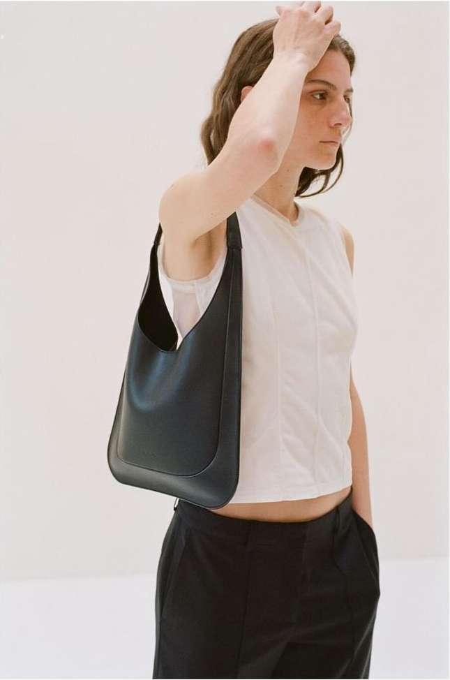 A woman with medium-length brown hair poses indoors with a black leather shoulder bag, wearing a white sleeveless top and black pants.