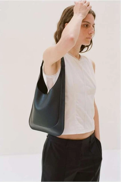 A woman with medium-length brown hair poses indoors with a black leather shoulder bag, wearing a white sleeveless top and black pants.