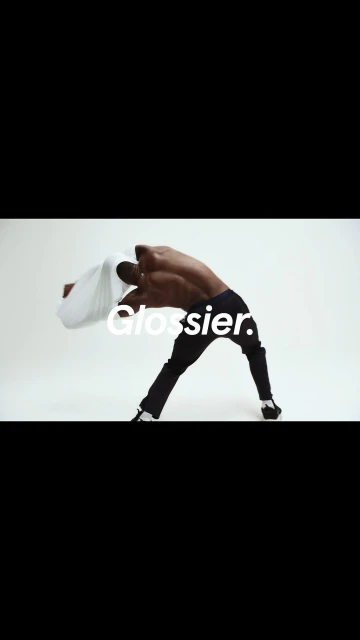 Shirtless man in black pants arches backwards, holding a white shirt against a white background, with the text "Glossier."