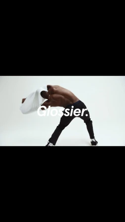 Shirtless man in black pants arches backwards, holding a white shirt against a white background, with the text "Glossier."