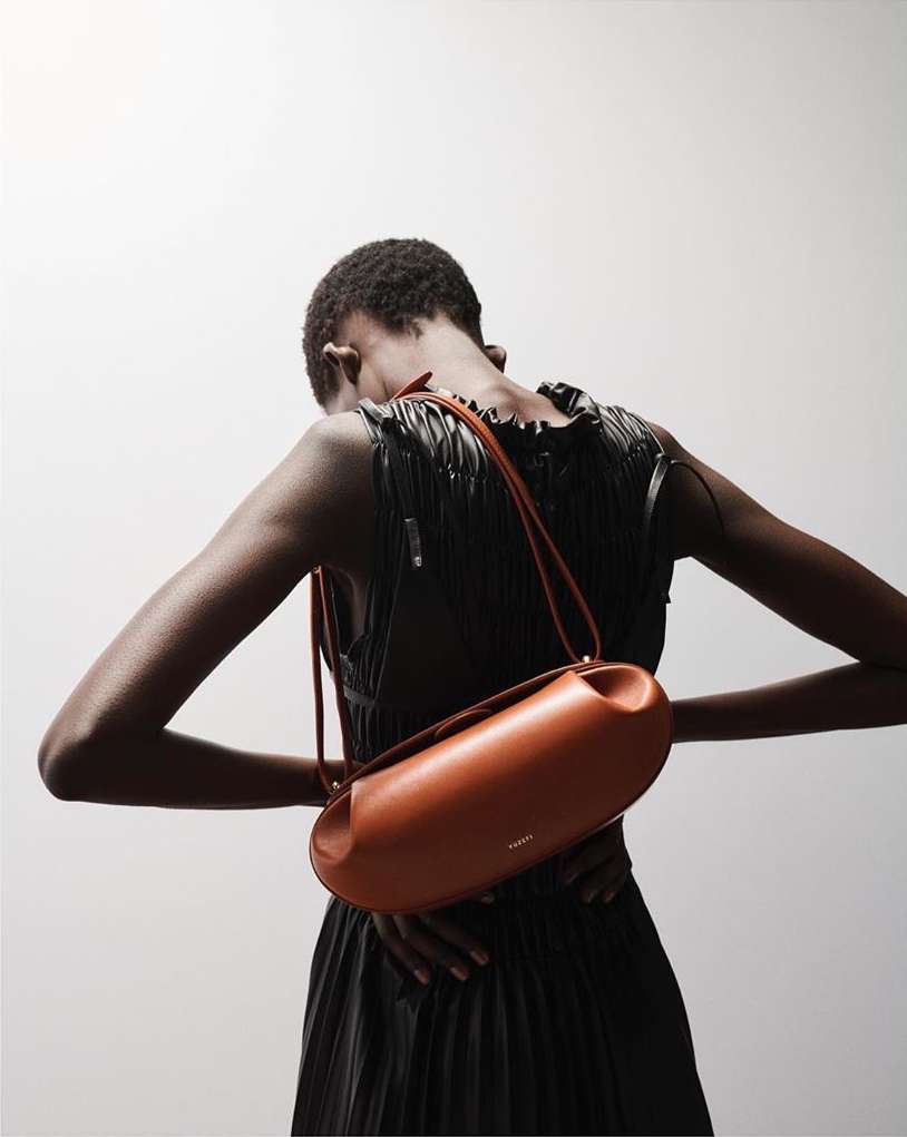Fashion shot featuring a slender Black woman with a tan leather purse slung over her shoulder, wearing a black dress against a white backdrop.