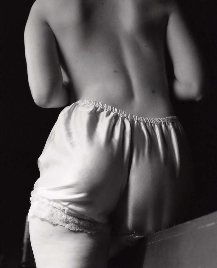 Monochrome shot from behind of an adult female with bare back and white lace-trimmed satin shorts against a dark background in soft lighting.