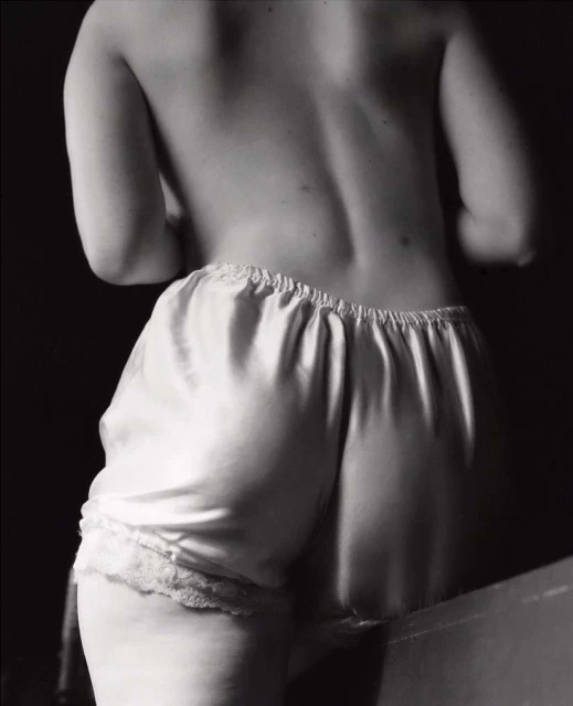 Monochrome shot from behind of an adult female with bare back and white lace-trimmed satin shorts against a dark background in soft lighting.