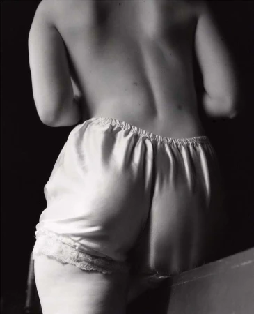 Monochrome shot from behind of an adult female with bare back and white lace-trimmed satin shorts against a dark background in soft lighting.