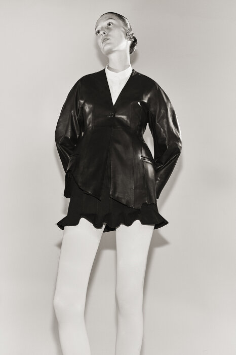 Model in black leather jacket and white tights against a plain light background photographed by Sarah Blais