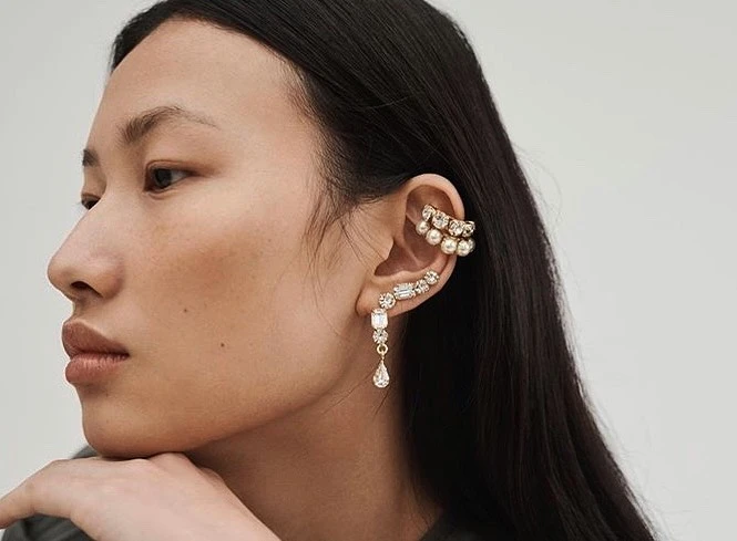 Profile of a woman with dark hair wearing pearl and crystal ear cuffs and drop earrings on a solid light background in a studio.