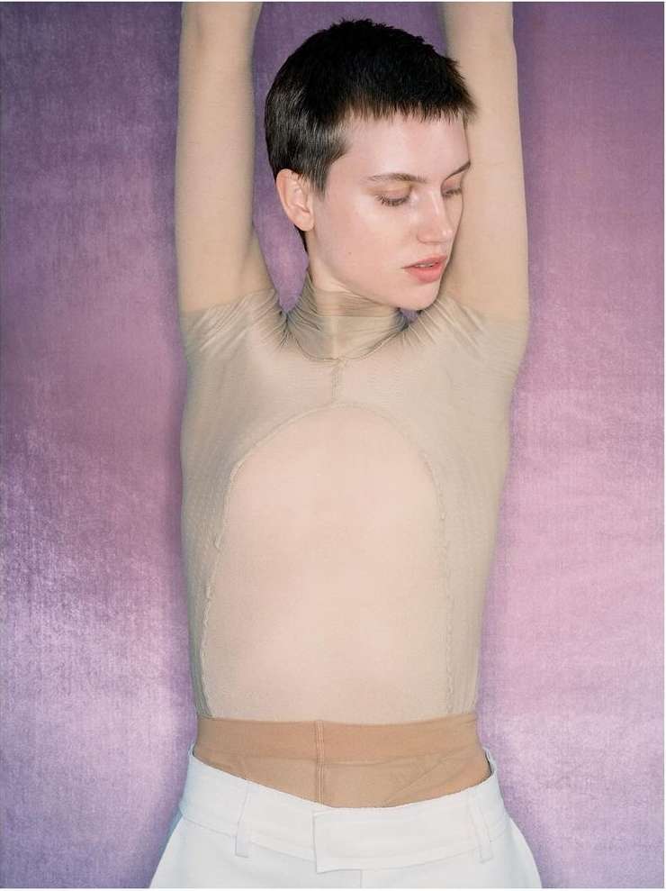 Portrait of a young adult woman with short hair posing in a sheer top and white pants against a textured lavender background.