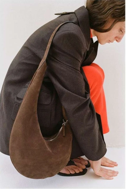 Fashion shot featuring a brunette woman in a charcoal coat and orange pants, posing with a brown suede hobo bag in a studio.