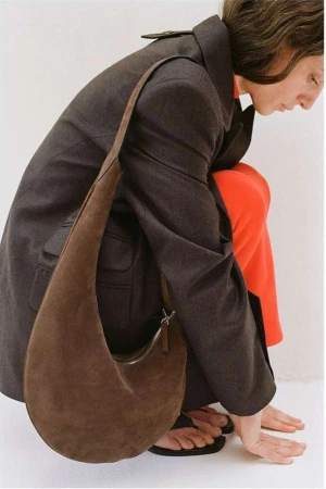 Fashion shot featuring a brunette woman in a charcoal coat and orange pants, posing with a brown suede hobo bag in a studio.