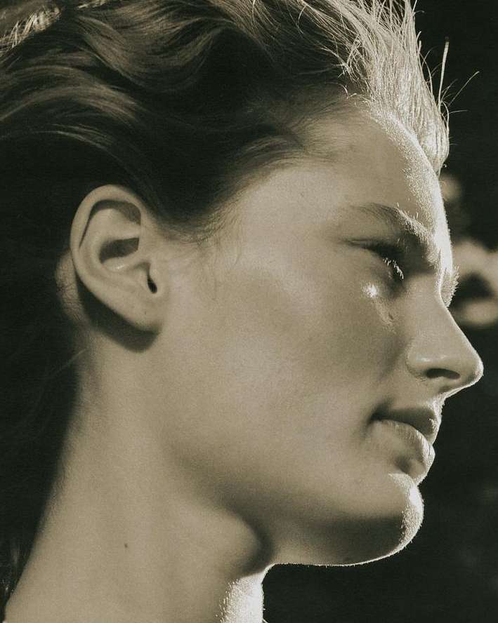 Monochrome profile portrait of an adult female with fair skin and dark hair, eyes closed, against a dark background, under bright lighting.