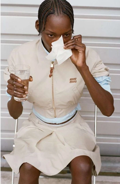 A woman with braided hair, wearing a tan jacket and skirt, sits on a chair holding a tissue to her nose and a glass of water.