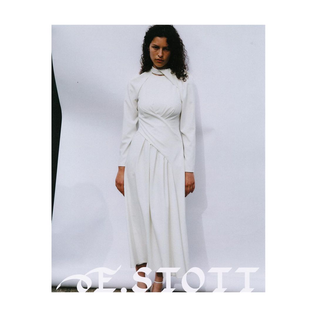 A woman in a long-sleeved, white dress with an asymmetrical neckline stands against a neutral backdrop in a fashion forward editorial setting.