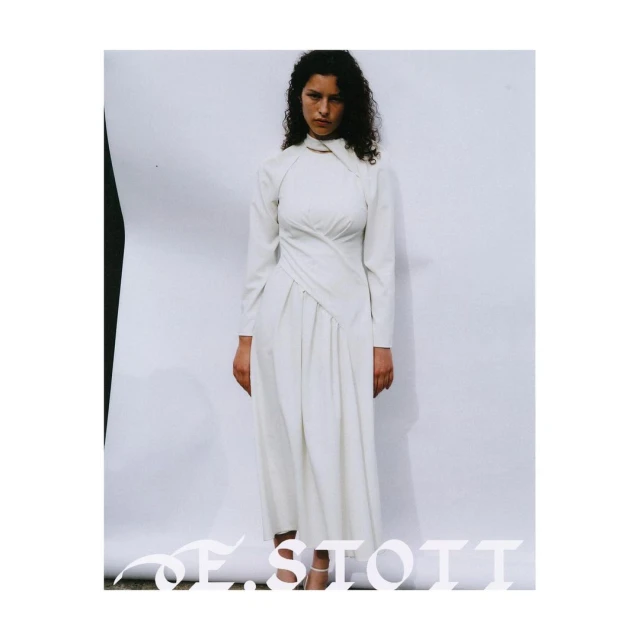 A woman in a long-sleeved, white dress with an asymmetrical neckline stands against a neutral backdrop in a fashion forward editorial setting.