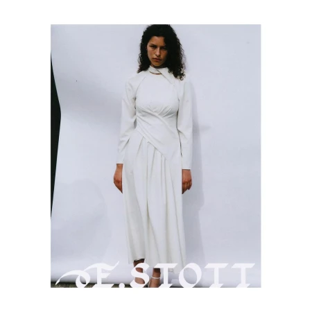 A woman in a long-sleeved, white dress with an asymmetrical neckline stands against a neutral backdrop in a fashion forward editorial setting.