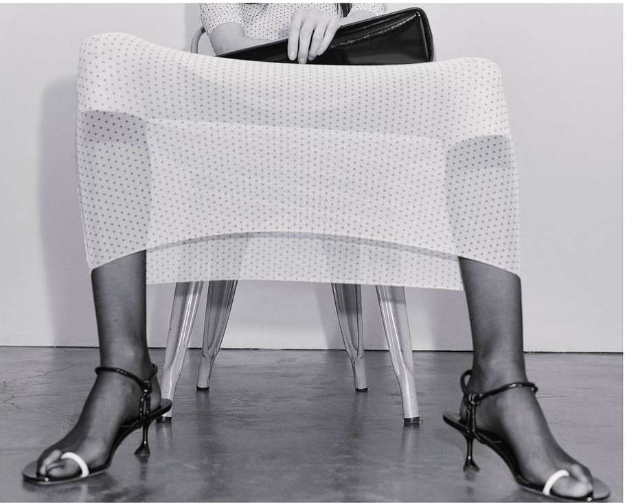 Fashion shot of a woman sitting with handbag, wearing polka dot dress, black hosiery, and black sandals with white toe straps.