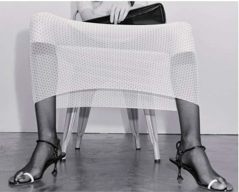Fashion shot of a woman sitting with handbag, wearing polka dot dress, black hosiery, and black sandals with white toe straps.
