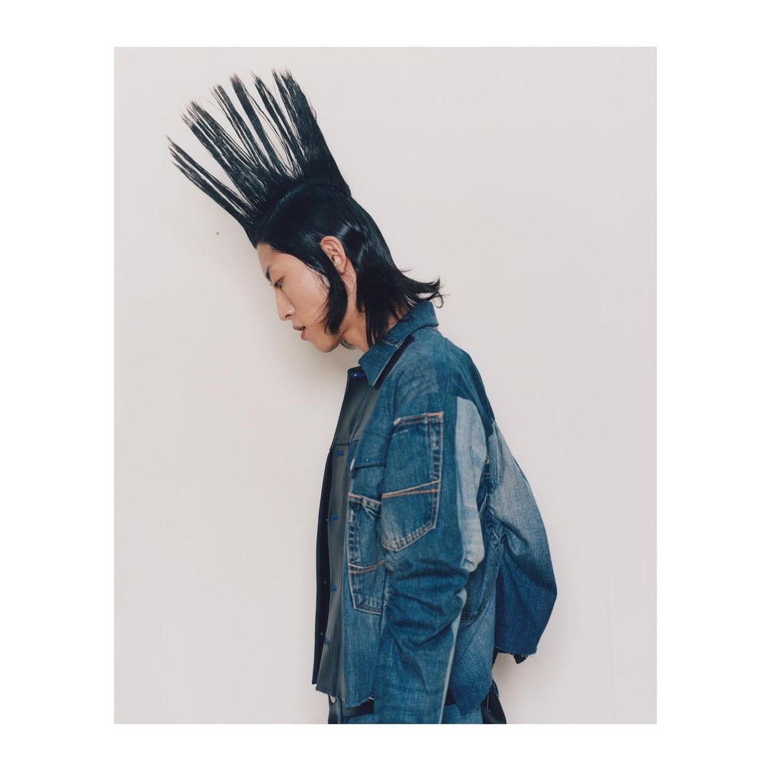 Profile of a stylish young adult with a spiky mohawk hairstyle and wearing a denim jacket against a plain white backdrop.