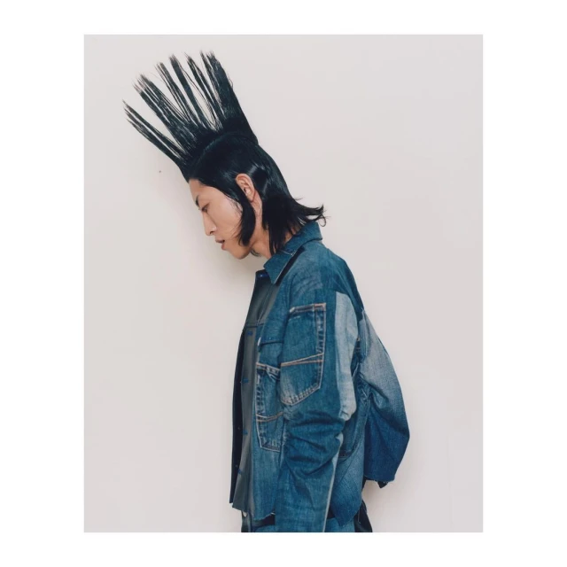 Profile of a stylish young adult with a spiky mohawk hairstyle and wearing a denim jacket against a plain white backdrop.