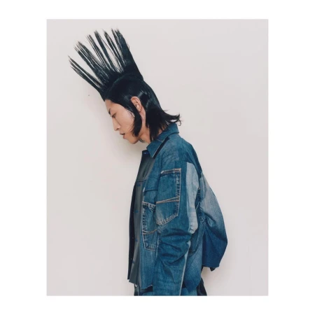 Profile of a stylish young adult with a spiky mohawk hairstyle and wearing a denim jacket against a plain white backdrop.