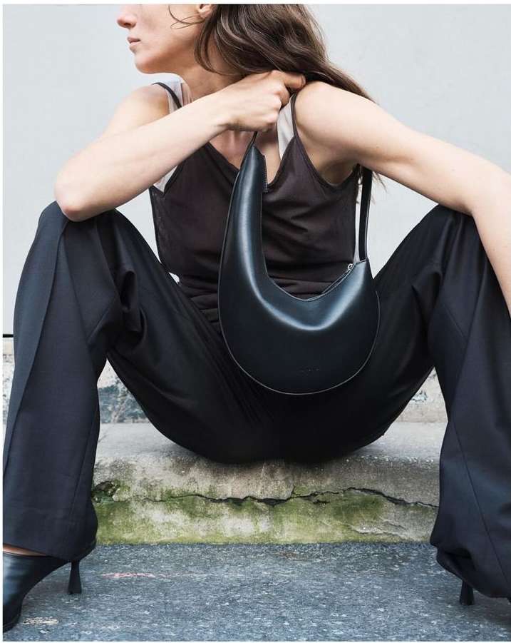Woman in black outfit with black crescent-shaped purse sitting on concrete steps, minimalist fashion editorial style in daylight.