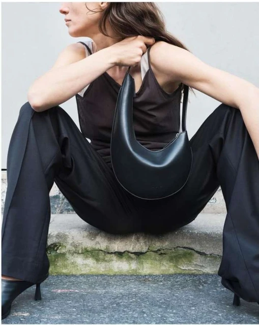 Woman in black outfit with black crescent-shaped purse sitting on concrete steps, minimalist fashion editorial style in daylight.