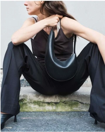 Woman in black outfit with black crescent-shaped purse sitting on concrete steps, minimalist fashion editorial style in daylight.