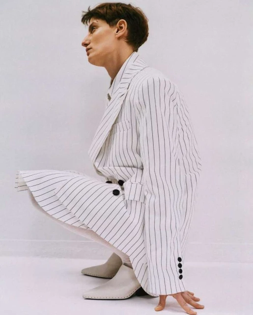 Person in white pinstripe suit crouching indoors, wearing light-colored ankle boots against a plain white wall and floor background.