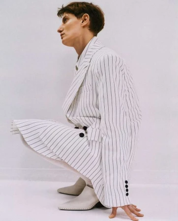 Person in white pinstripe suit crouching indoors, wearing light-colored ankle boots against a plain white wall and floor background.