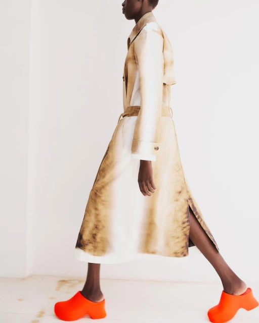 Fashion model in a beige trench coat walks with orange clogs in a studio. The shot is a medium shot, and the background is white.