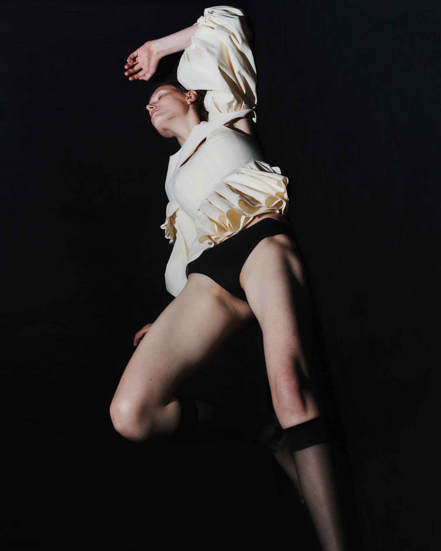 Low-angle shot of a solo female model in a light top and black bottoms striking a pose against a dark backdrop in a studio setting.