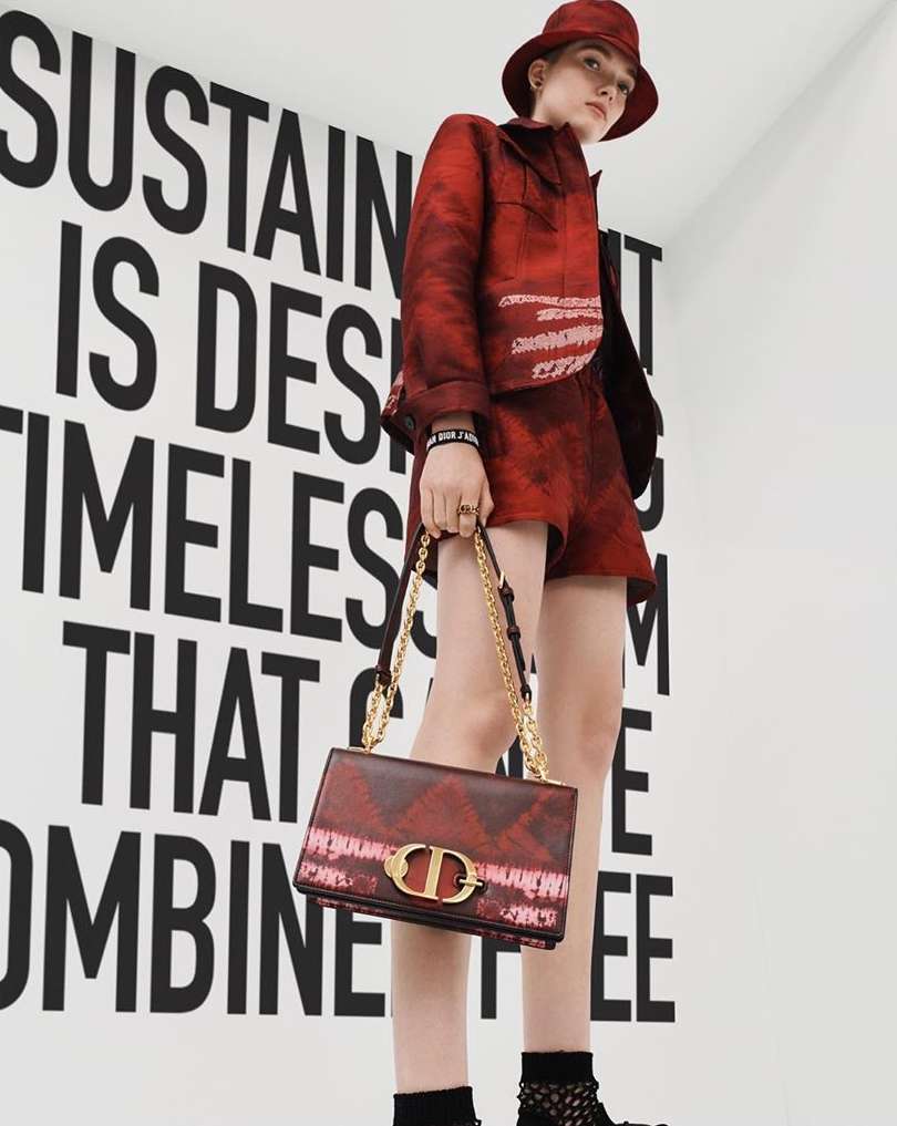 A fashion model poses indoors next to a large typographical display wearing a matching red jacket, shorts, hat, and carrying a handbag.