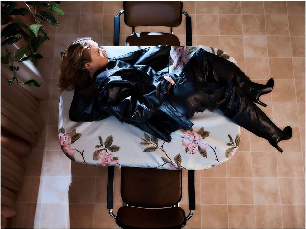 Woman in black leather jacket and thigh-high boots reclines on a floral-patterned table, flanked by chairs in an indoor setting.