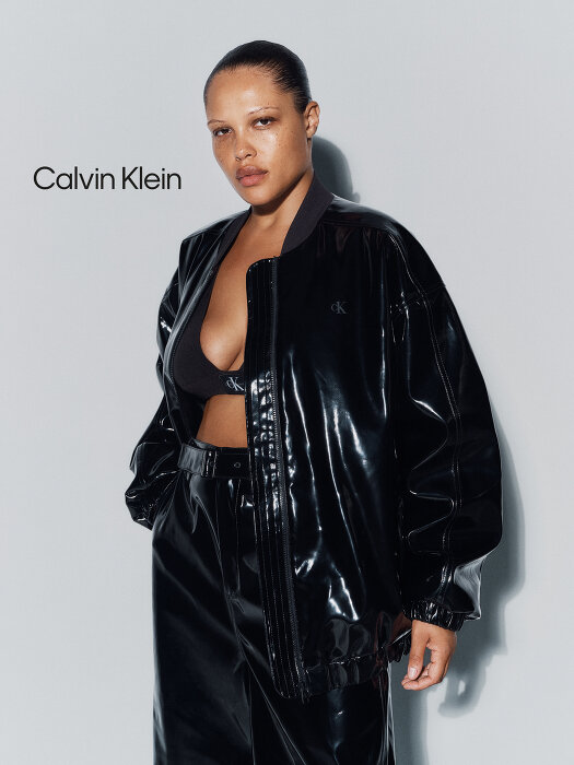 Model wearing a glossy black vinyl jacket and matching trousers against a pale grey studio background.