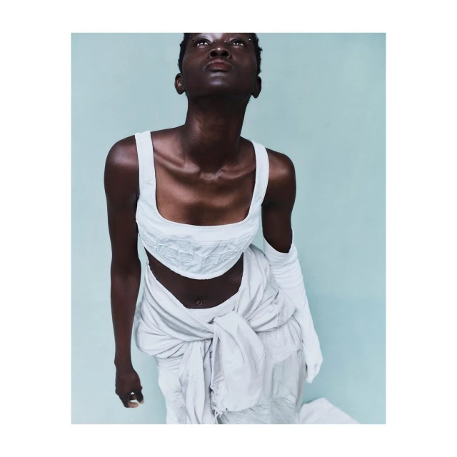 African-American female fashion model poses in studio with off-white crop top, skirt tied at waist, long gloves, and upward gaze.