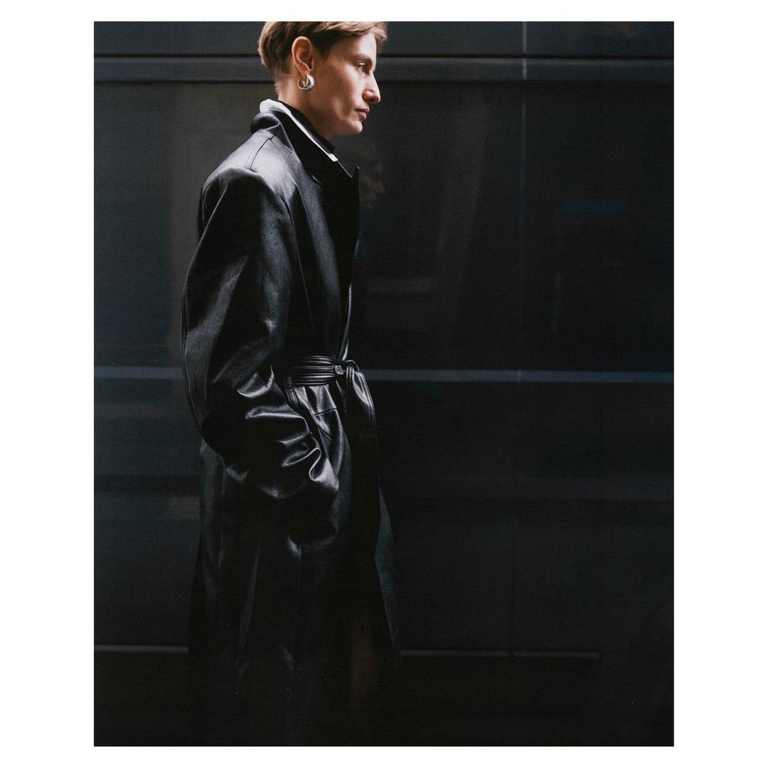 Side profile of a person with short hair wearing a long black leather coat against a dark grey paneled wall, fashion-forward style.