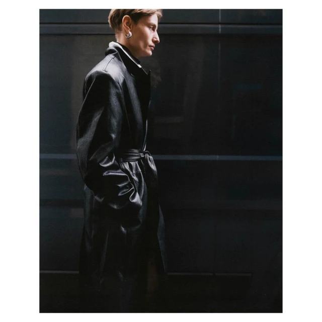 Side profile of a person with short hair wearing a long black leather coat against a dark grey paneled wall, fashion-forward style.