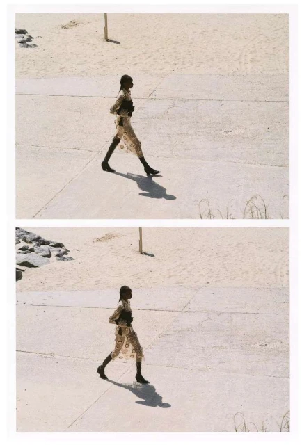 Fashionable woman in patterned dress walks on concrete in an outdoor scene, with neutral sand, grass, and distant rocks in the background.