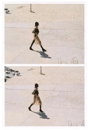 Fashionable woman in patterned dress walks on concrete in an outdoor scene, with neutral sand, grass, and distant rocks in the background.