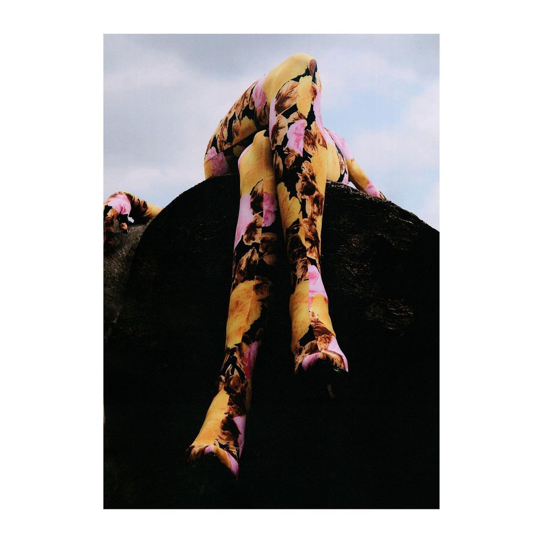 A person wearing a floral pattern sits atop a rock against a partly cloudy sky in a fashion-forward outdoor shot with dark shadows.