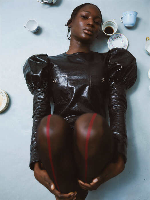 Striking studio portrait of a Black woman in a glossy dark dress holding her knees, accessorized with teacups on a grey background.