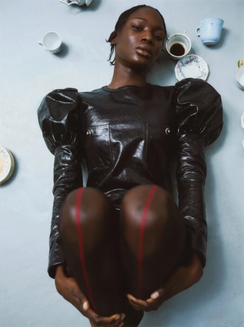 Striking studio portrait of a Black woman in a glossy dark dress holding her knees, accessorized with teacups on a grey background.